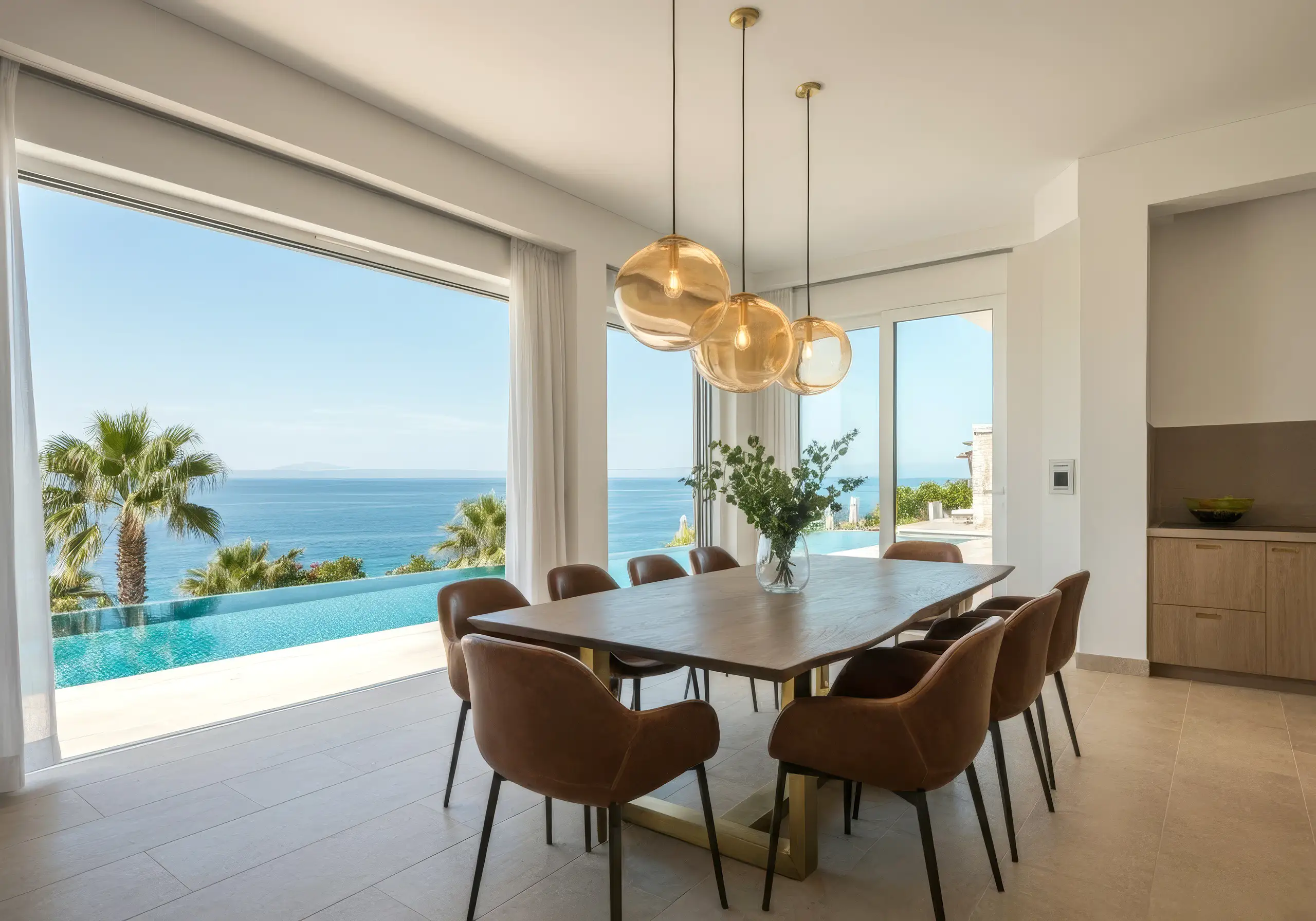 Dining table with brown chairs and glass pendants before a glass wall onto the pool and ocean
