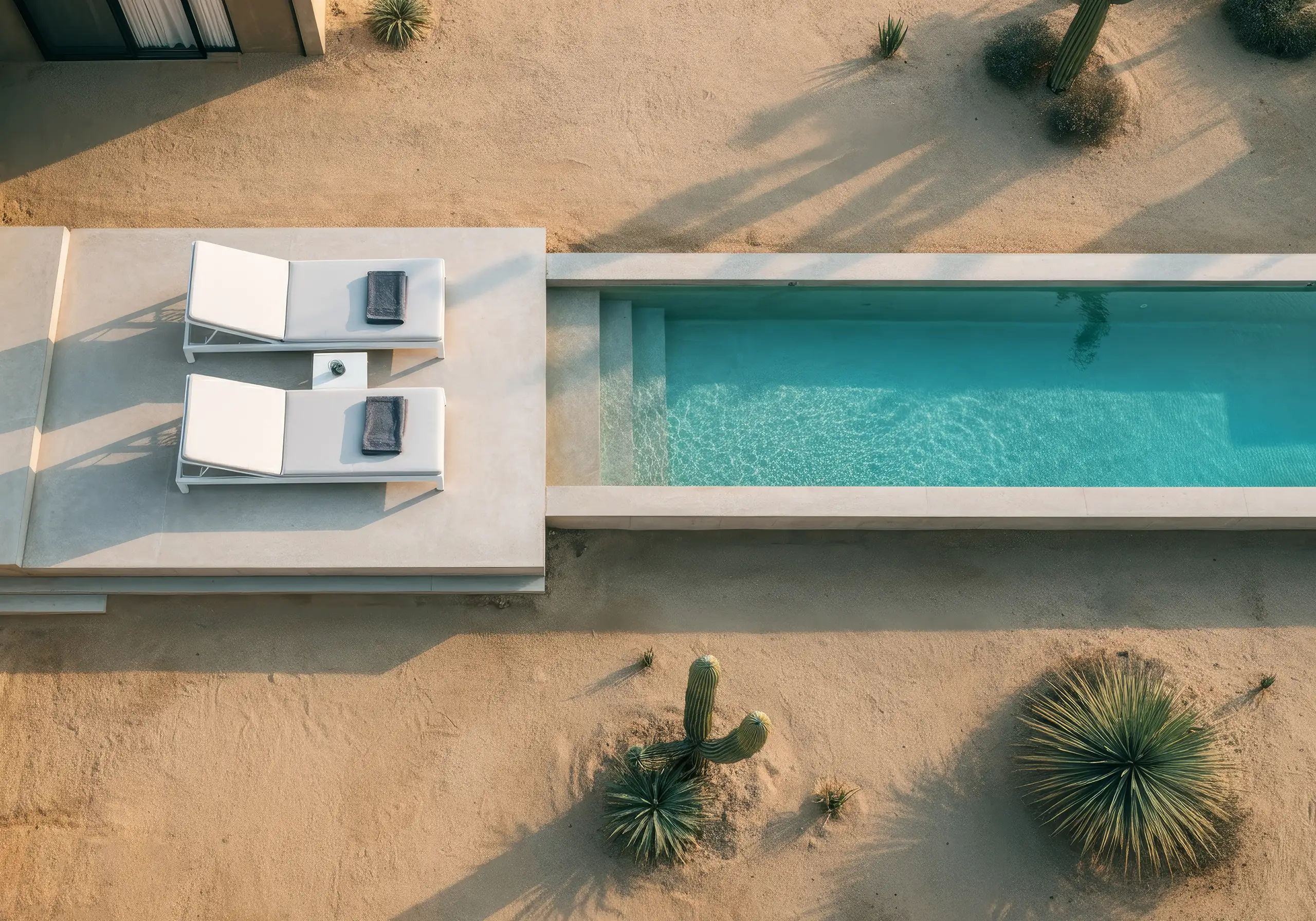 Aerial view of a narrow pool and white loungers set in desert sand and cacti