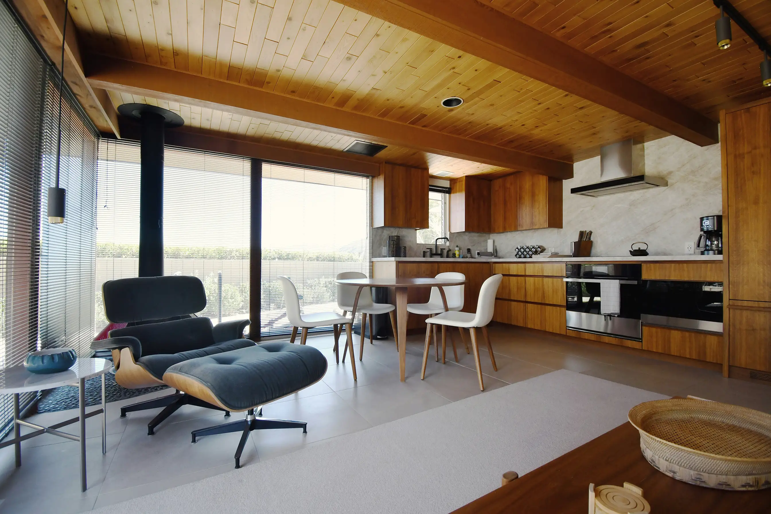 Open living area with a lounge chair, a wood stove and a wooden kitchen under a timber ceiling