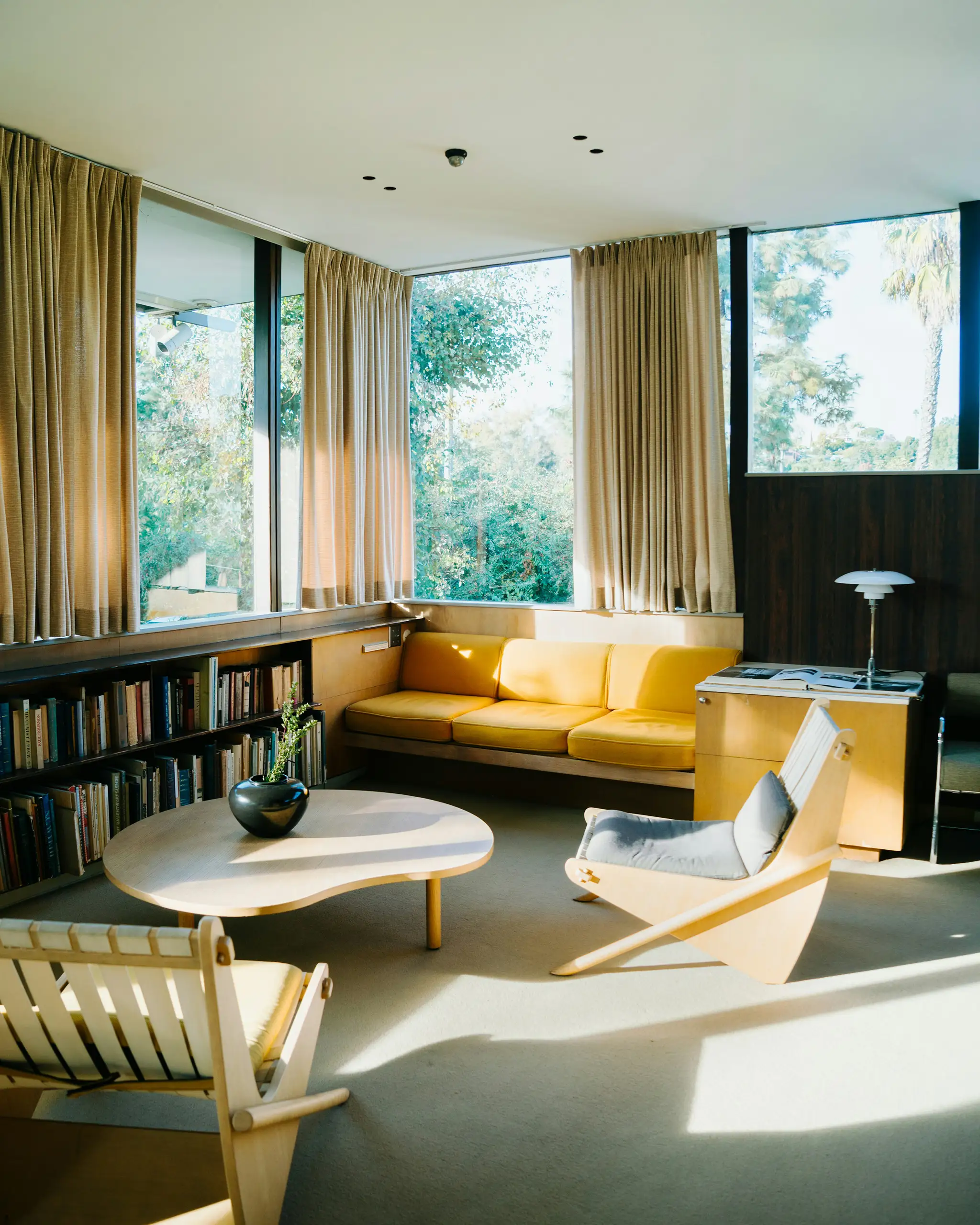Corner living room with a yellow sofa, bookshelves and windows onto the trees