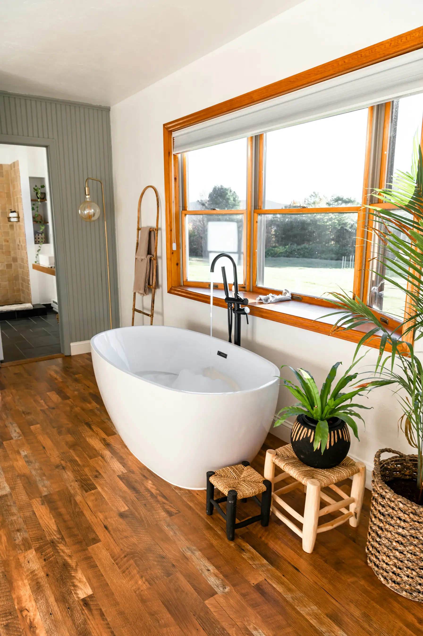 Bathroom with a freestanding tub beneath a wood-framed window and a wooden floor
